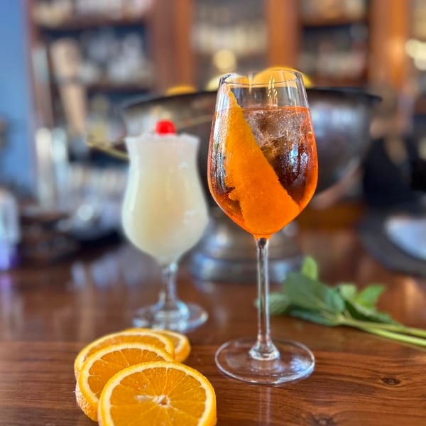 Two cocktails, one orange with an orange peel and one white with a cherry, on a wooden bar.