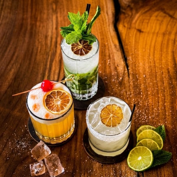 Three vibrant cocktails, including a mojito, on a rustic wooden table with lime slices and ice.