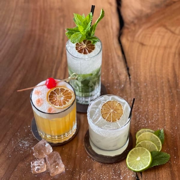 Three colorful cocktails with garnishes on a rustic wooden table with scattered sugar and lime slices.