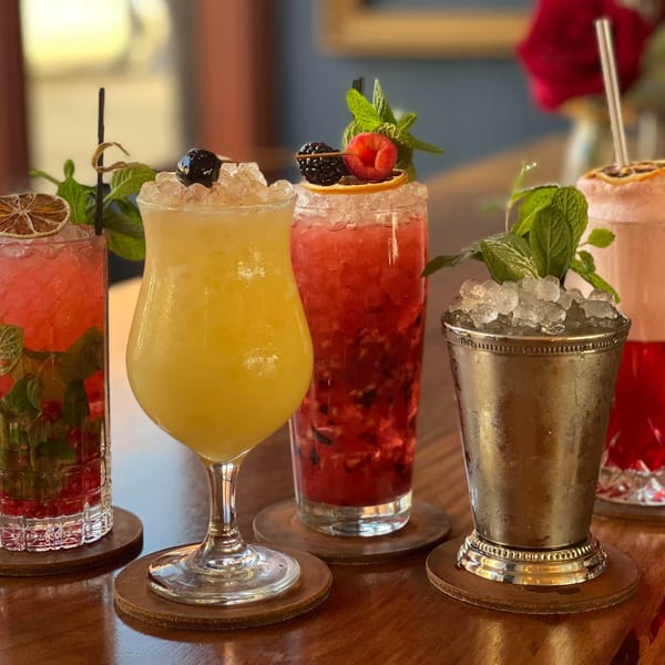 Five vibrant cocktails, garnished with fruit and mint, sit on a polished wooden table.
