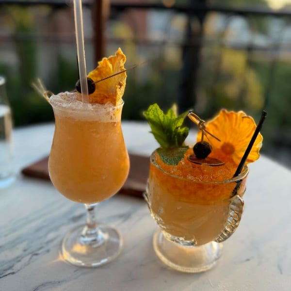 Two vibrant orange cocktails, garnished with pineapple, cherries, and mint, on a white marble table.